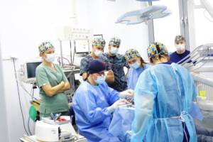 Fotografia z nowoczesnej sali zabiegowej w klinice CZAR-DENT, wykonana podczas szkolenia z zakresu zaawansowanej chirurgii stomatologicznej. W centrum kadru znajduje się dr n. med. Michał Czarnowski, który w lupach operacyjnych i sterylnym fartuchu z najwyższą precyzją przeprowadza zabieg u Pacjenta. Wokół fotela zgromadzona jest grupa skupionych lekarzy, którzy uważnie obserwują technikę i protokoły pracy eksperta. Zdjęcie doskonale ilustruje proces dzielenia się specjalistyczną wiedzą implantologiczną oraz pełne skupienie zespołu na komforcie i bezpieczeństwie leczonego Pacjenta.